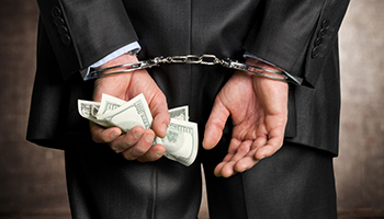 A man in a suit handcuffed and with some bills in one of their hands, representing white collar crimes